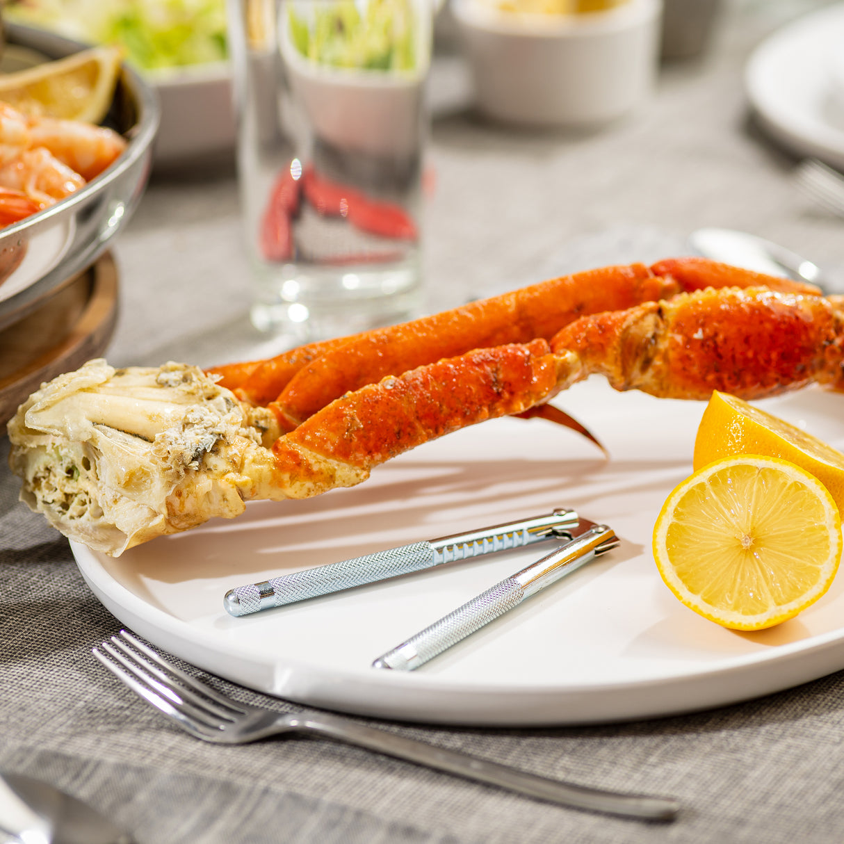 A plate with two large crab legs, a halved lemon, and stainless steel seafood crackers is set on a table with a gray cloth. In the background are blurred dishes, a glass of water, and utensils, suggesting a seafood meal.