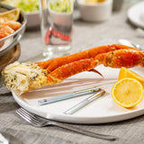A plate with two large crab legs, a halved lemon, and stainless steel seafood crackers is set on a table with a gray cloth. In the background are blurred dishes, a glass of water, and utensils, suggesting a seafood meal.