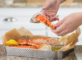 A person uses a chrome plated seafood cracker to open a red and white crab leg over a basket lined with parchment paper. Inside the basket are more crab legs, lemon wedges, and a glass dish of butter sauce on a granite counter.