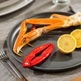 A black plate holds a crab leg, a red enamel cast aluminum cracker, and two lemon halves, all arranged on a gray placemat next to a fork. The scene suggests preparation for enjoying seafood with the perfect lobster crack tool.