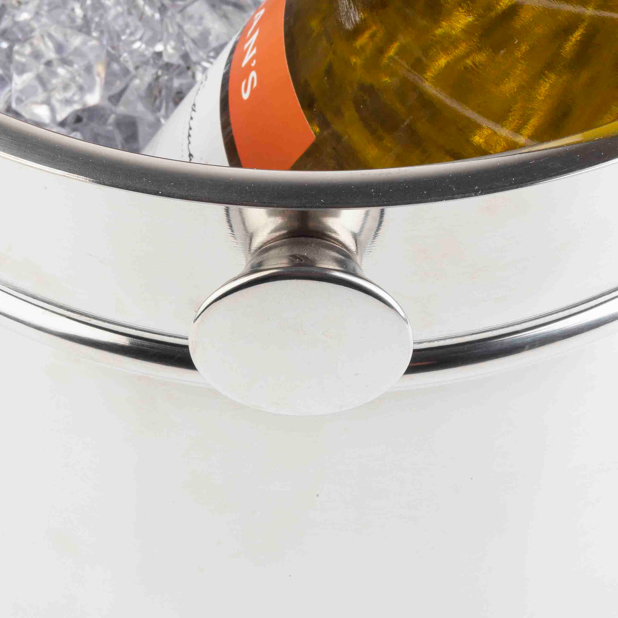 A close-up of a stainless steel wine/champagne bucket filled with ice cubes, holding a partially visible wine bottle with an orange label. The round handle of this dishwasher safe bucket is prominent at the center of the image.