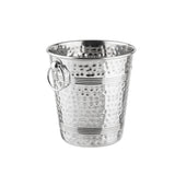 A shiny, stainless steel ice bucket with a hammered texture and horizontal grooves. Featuring a circular handle attached to a ring on its side, this TableCraft bucket is empty and placed against a plain white background.