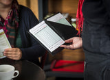 A server presents a black TableCraft bill folder with a receipt to a seated customer. The customer, wearing a colorful scarf and dark sweater, holds a menu near a white cup and saucer in this cozy restaurant setting.