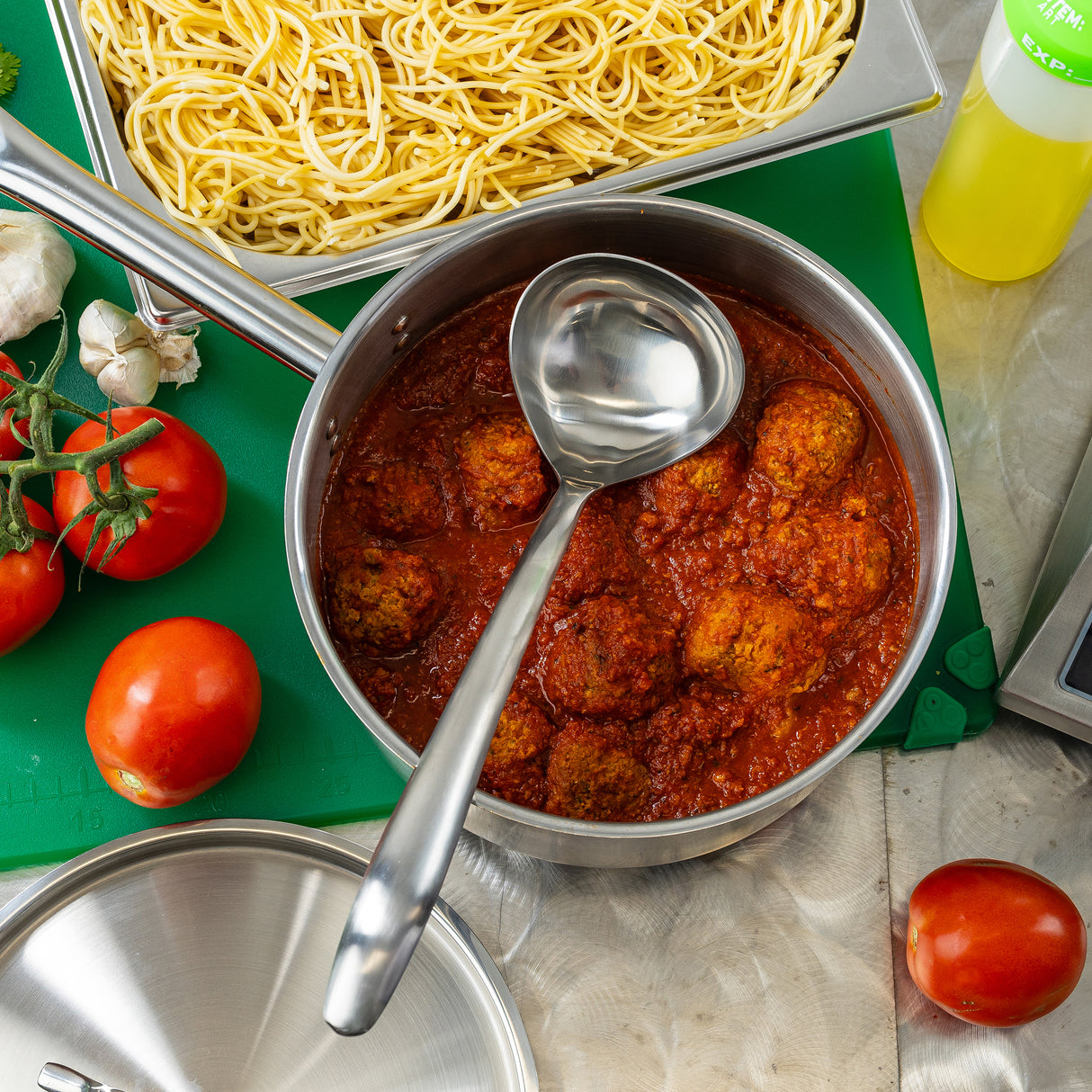 A Dalton Collection Soup Ladle rests in the pot of meatballs in red tomato sauce beside plain spaghetti. Nearby, fresh tomatoes, garlic, oil, and a metal lid sit on a green board. The ladle is dishwasher safe for easy cleanup.