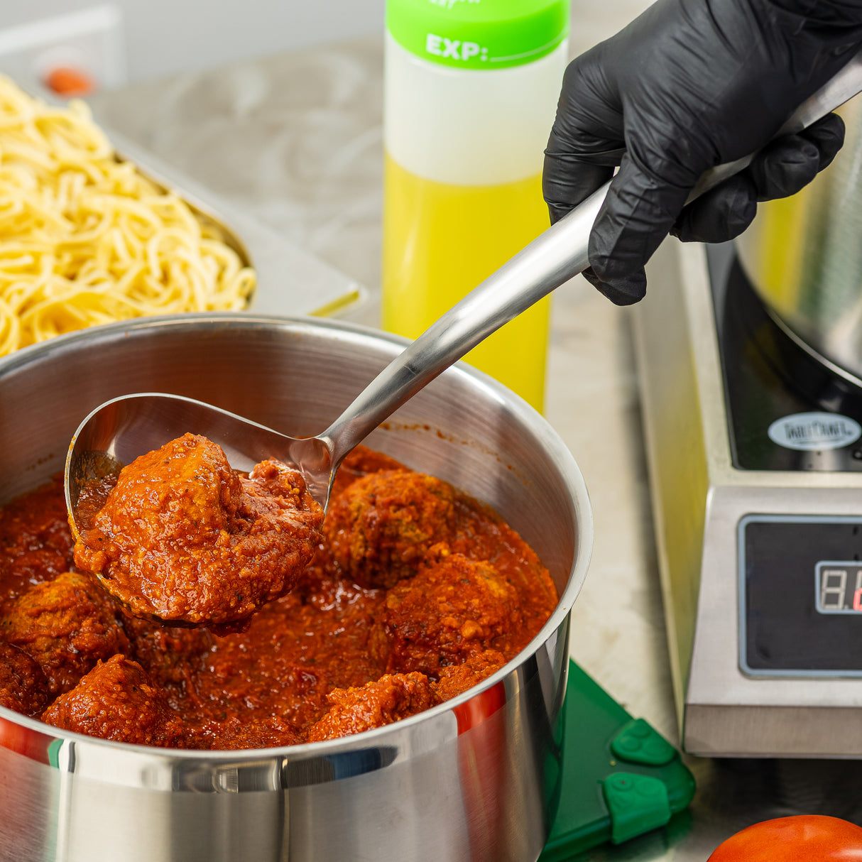A gloved hand holds a Dalton Collection Soup Ladle scooping meatballs in red tomato sauce from a stainless steel pot. In the background, uncooked pasta sits on a tray and a yellow squeeze bottle is visible next to a digital hot plate.