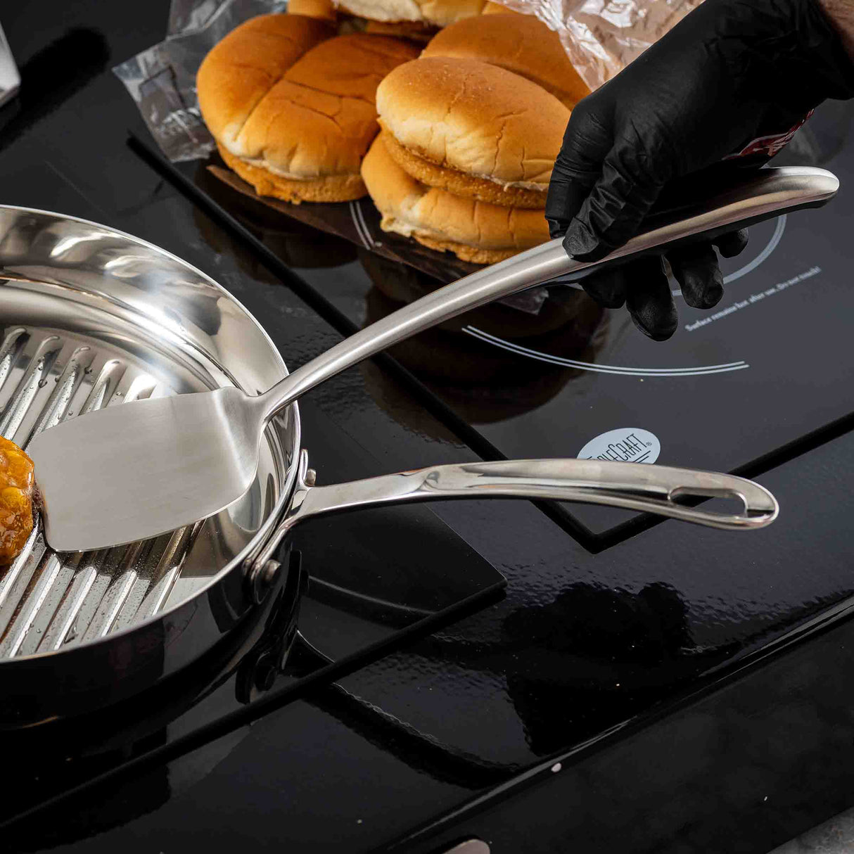 A person wearing a black glove uses a Dalton Collection Turner to cook a burger patty in a grill pan on the stovetop. Three hamburger buns rest on the counter in the background, partially in an open plastic bag, reflecting a kitchen setting.
