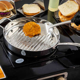 A hand in a black glove uses a Dalton Collection Turner to lift a cooked burger patty with melted cheese from a stainless steel grill pan. Open buns and toppings like lettuce, onion, and tomato are arranged nearby, ready for assembling.