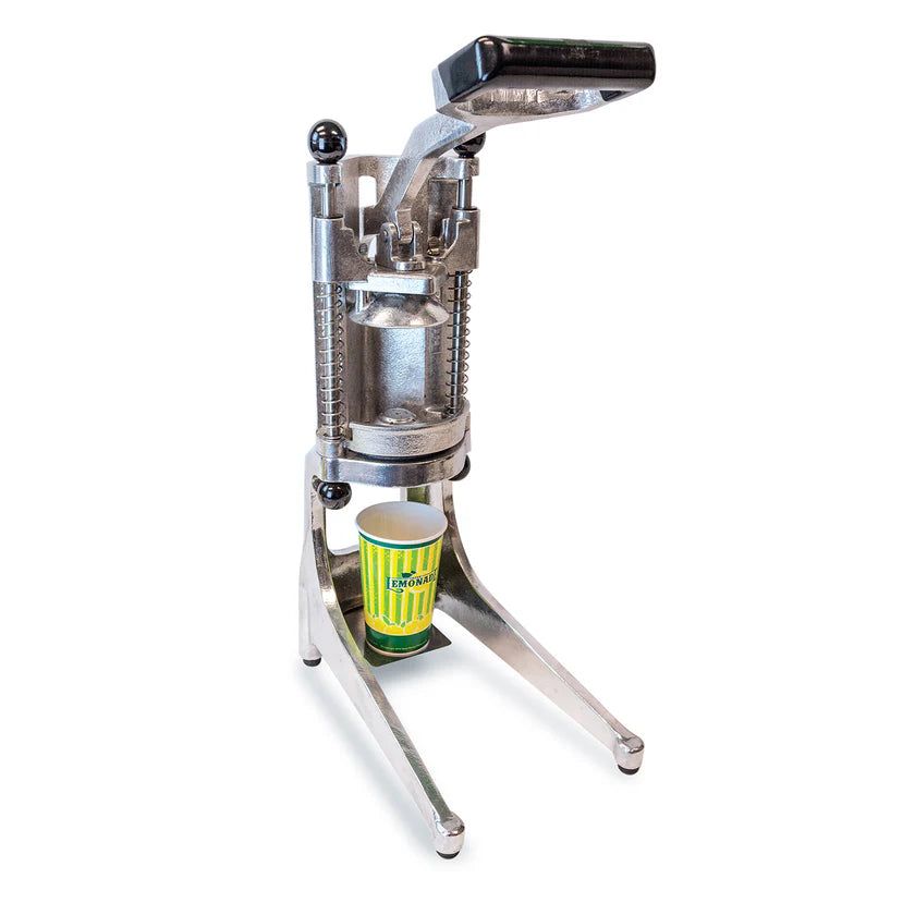 A metal manual Lemon Squeezer stands on a white background. Its handle is raised, and a yellow-green striped paper cup labeled Lemon is positioned underneath, ready to catch juice. The aluminum device has sturdy legs and visible springs.
