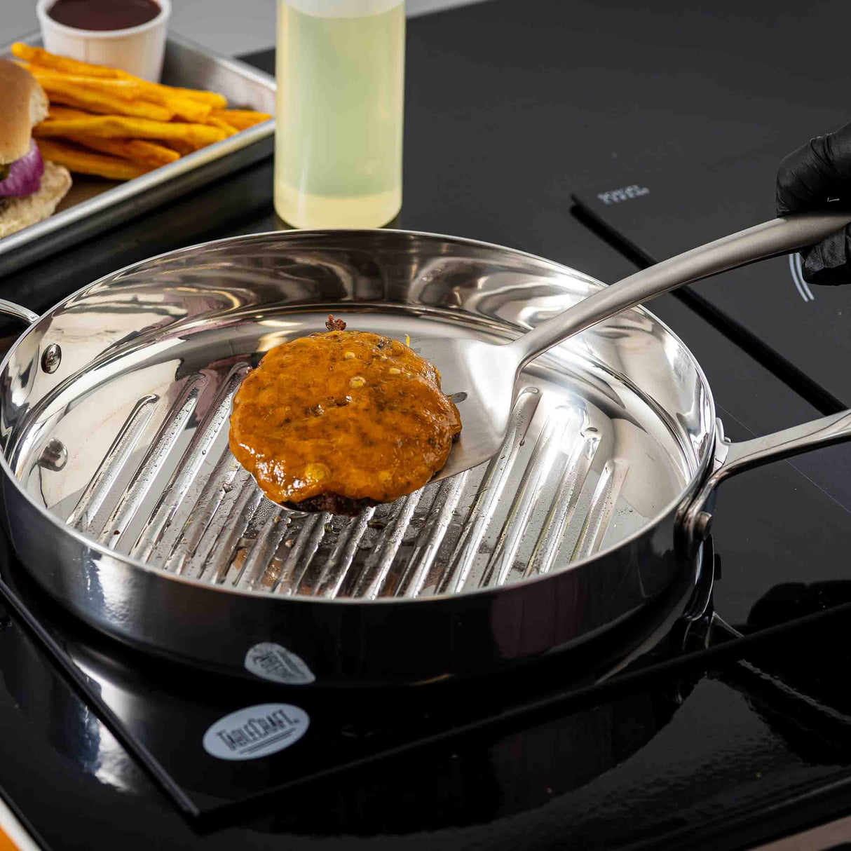 A gloved hand uses a Dalton Collection Turner to cook a sauced burger patty on a stainless steel grill pan atop a black stovetop. In the background, fries, a burger, sauce, and a bottle of oil are visible.