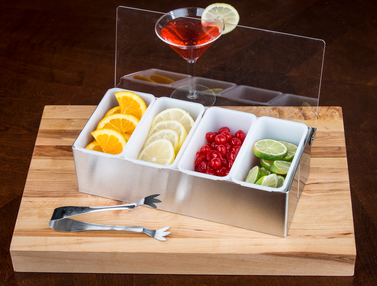 A stainless steel condiment holder with four compartments—filled with orange slices, lemon slices, maraschino cherries, and lime wedges—rests on a wooden board. Pom Tongs lie beside it, and a red cocktail with a lime wheel sits atop the holder.