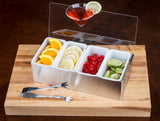 A stainless steel condiment holder with four compartments—filled with orange slices, lemon slices, maraschino cherries, and lime wedges—rests on a wooden board. Pom Tongs lie beside it, and a red cocktail with a lime wheel sits atop the holder.