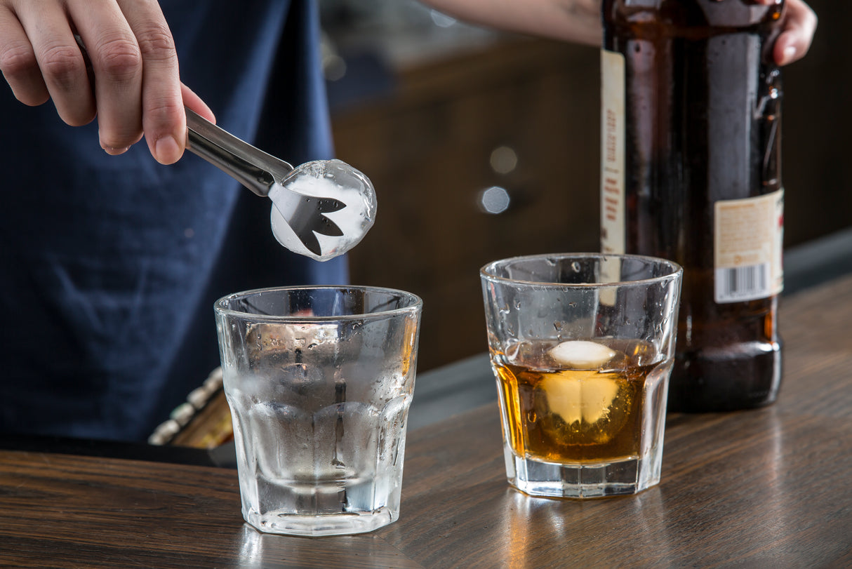 A person uses stainless steel Pom Tongs to place an ice cube into an empty glass. Next to it, a glass of brown liquor with ice and an amber bottle rest on a wooden surface, evoking a cozy bar or home setting. Pom Tongs are dishwasher safe for easy cleaning.