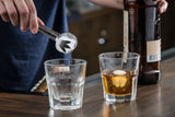 A person uses stainless steel Pom Tongs to place an ice cube into an empty glass. Next to it, a glass of brown liquor with ice and an amber bottle rest on a wooden surface, evoking a cozy bar or home setting. Pom Tongs are dishwasher safe for easy cleaning.