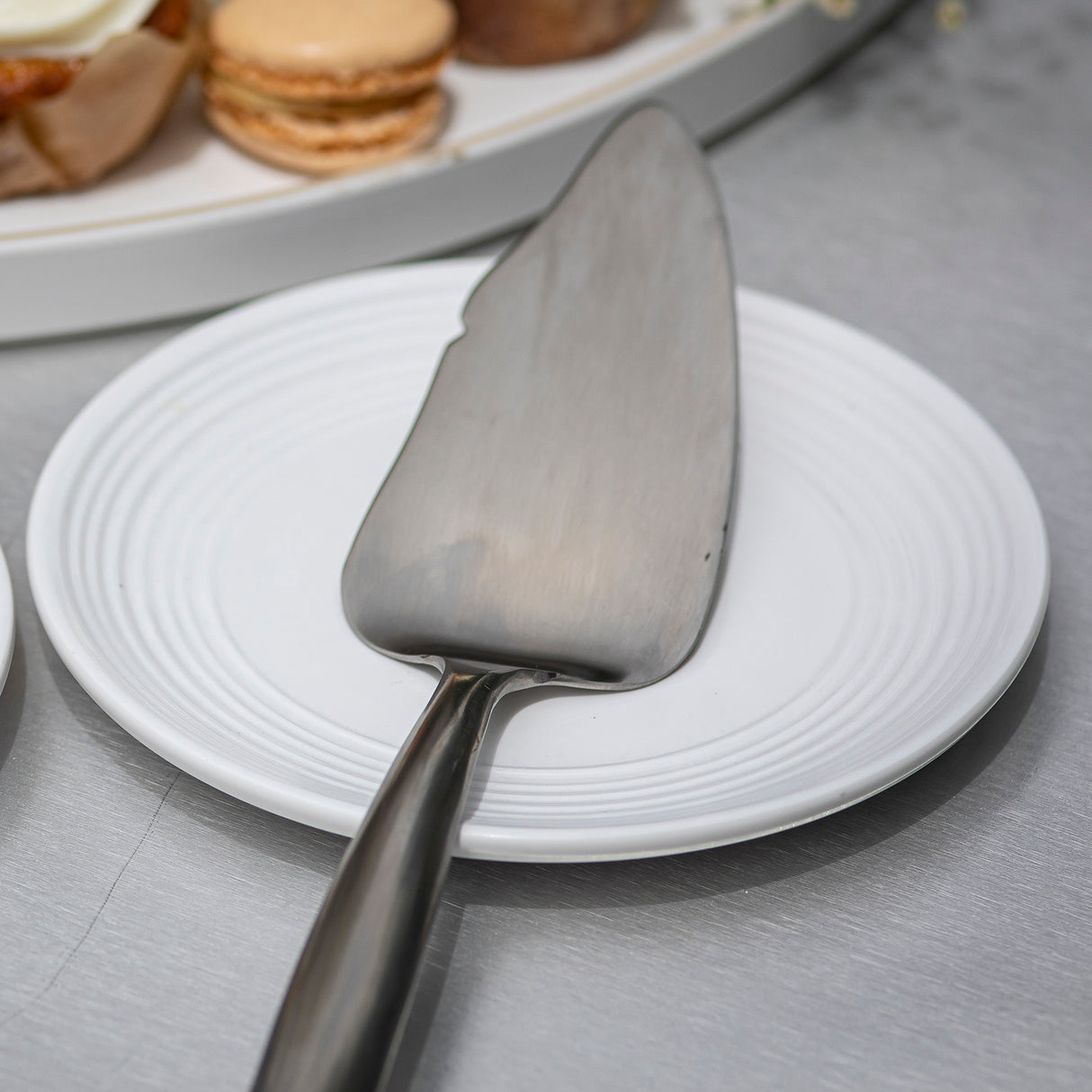 A Dalton Collection Buffet Cake Server rests on a small, round white plate with concentric circle patterns. In the background, a tray holds macarons and assorted pastries, slightly out of focus on a smooth metallic surface.
