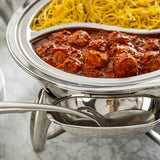 A round stainless steel serving dish contains spaghetti on one side and meatballs in red tomato sauce on the other. A Dalton Collection serving spoon, which is dishwasher safe, rests on the dish’s holder atop a marble countertop.