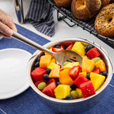 A hand holds a Dalton Collection Buffet Spoon over a metal bowl filled with colorful fruit salad, including strawberries, watermelon, pineapple, and more. Bagels and a stack of white plates sit nearby on a blue cloth.