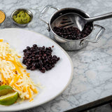 A white plate holds shredded yellow and white cheese, a wedge of lime, and black beans. Nearby, a small pot with a Dalton Collection Buffet Spoon contains more black beans. Small bowls of green and creamy brown sauces sit on the marble surface.