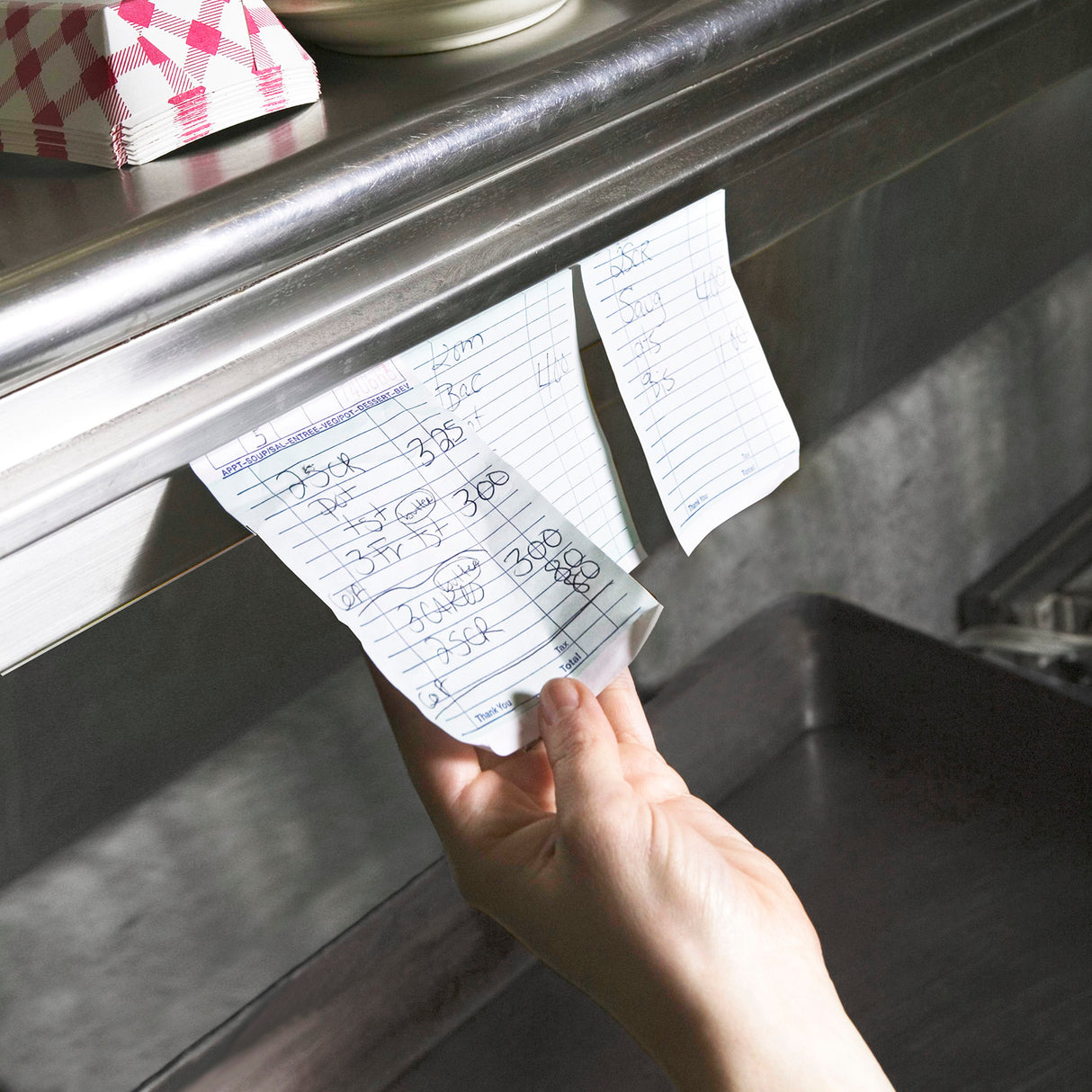 A hand pulls a handwritten restaurant order ticket from an aluminum order rack above a kitchen counter. Additional order tickets and pink-and-white checkered paper food trays fill the background, highlighting the busy kitchen environment.