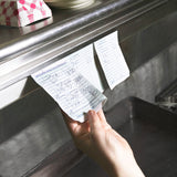 A hand pulls a handwritten restaurant order ticket from an aluminum order rack above a kitchen counter. Additional order tickets and pink-and-white checkered paper food trays fill the background, highlighting the busy kitchen environment.