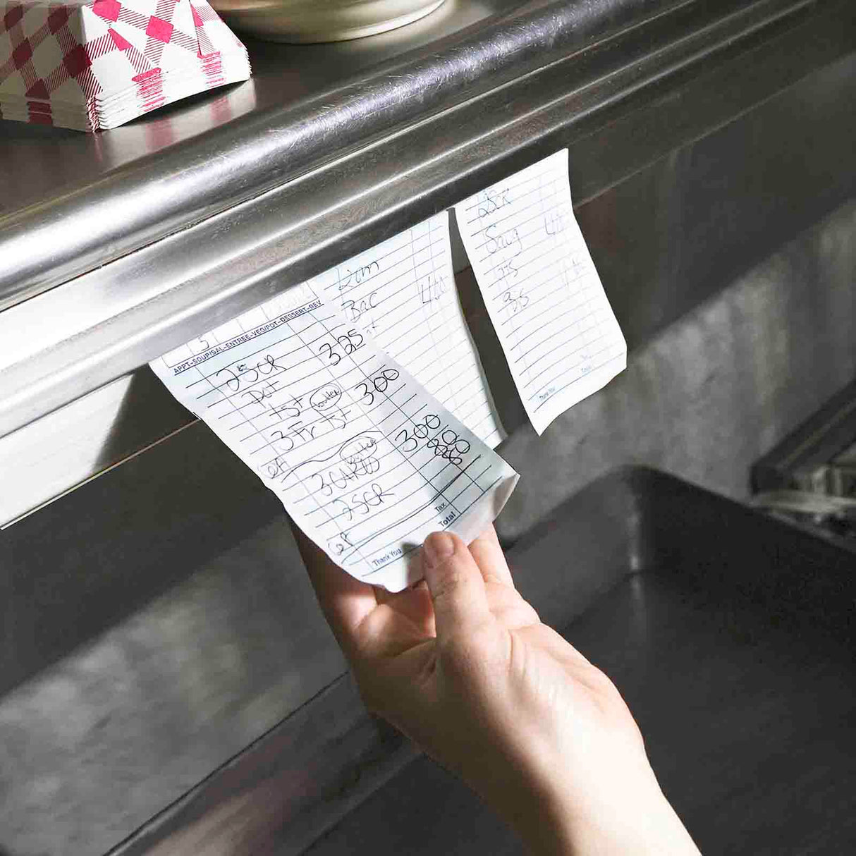 A hand pulls a paper order ticket from an aluminum order rack in a restaurant kitchen. Another ticket remains on the rail. Both tickets have handwritten food orders. A stack of napkins and a bowl are visible on the brightly lit shelf above.