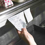 A hand pulls a paper order ticket from an aluminum order rack in a restaurant kitchen. Another ticket remains on the rail. Both tickets have handwritten food orders. A stack of napkins and a bowl are visible on the brightly lit shelf above.