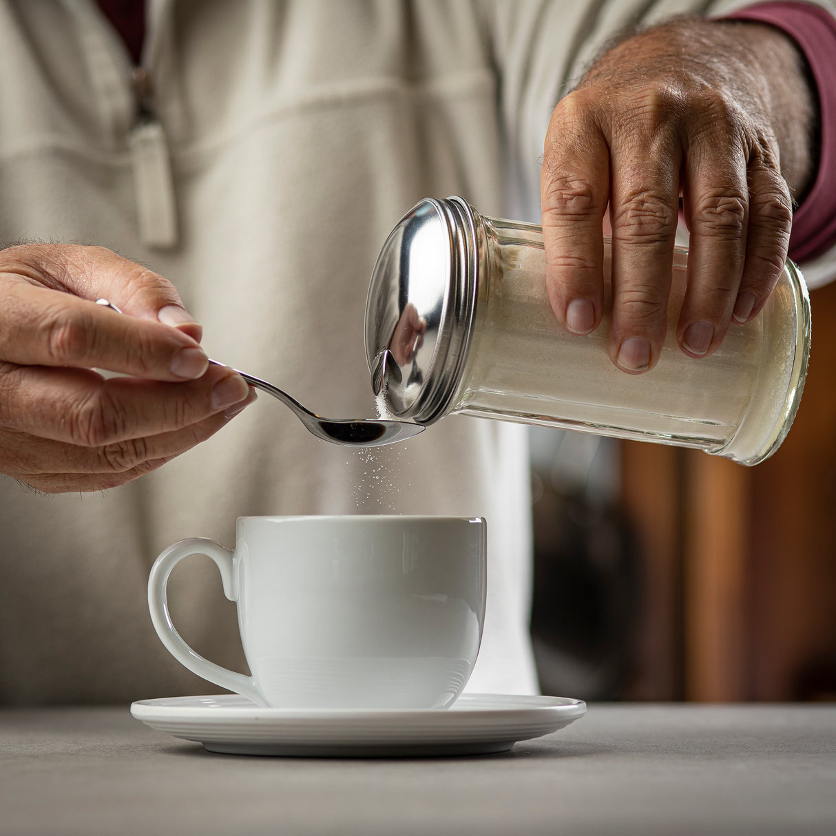 A person pours sugar from a 12 oz glass dispenser onto a spoon held over a white cup and saucer. The hands are close up, and the background is softly blurred, keeping focus on the action and the dishwasher safe items on the table.