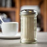 A fluted glass sugar pourer with a metal lid sits on a counter, filled with granulated sugar. In the blurred background, there is a white coffee cup and saucer. The dispenser is dishwasher safe, perfect for any kitchen or café setting.