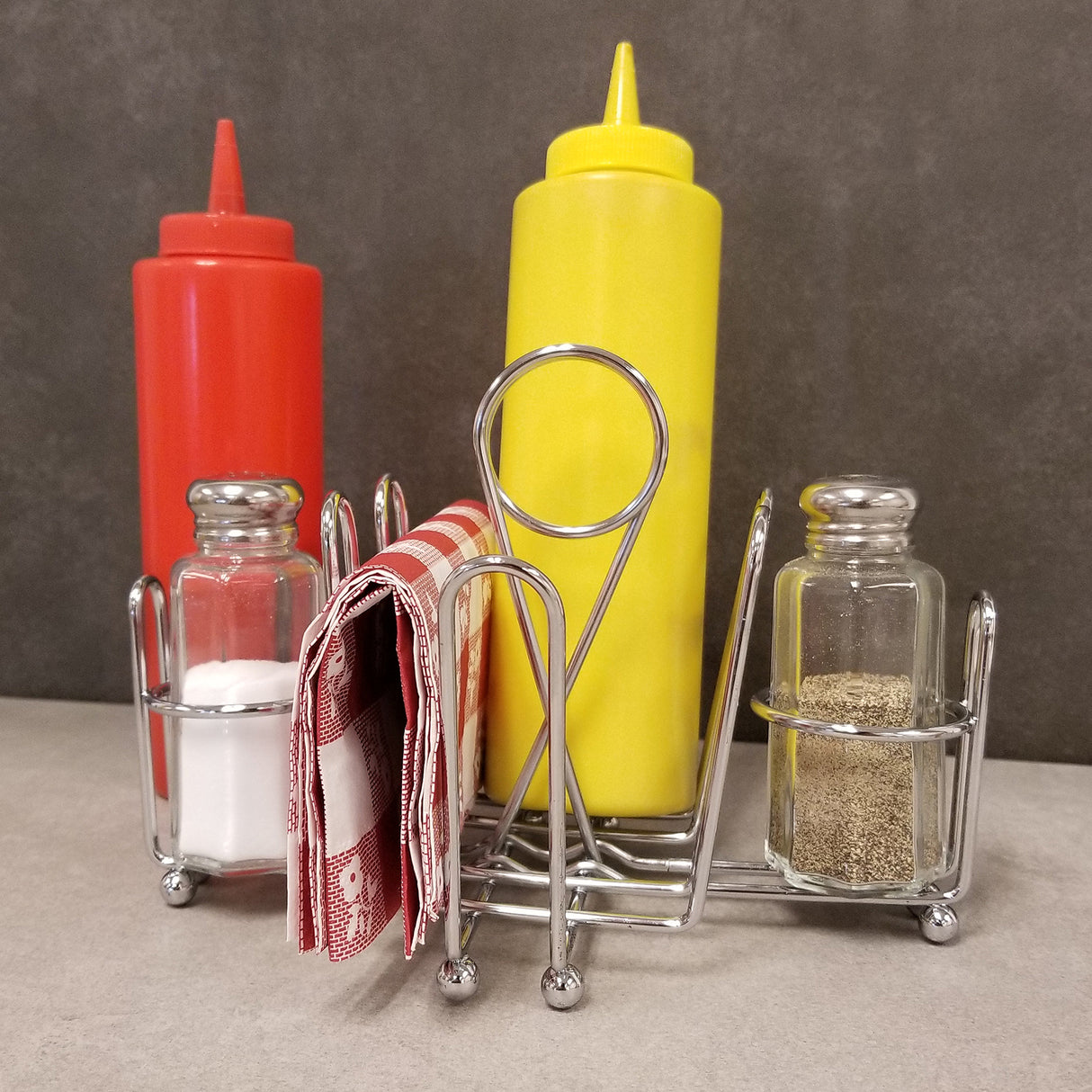A chrome plated combination rack holds a red ketchup bottle, a yellow mustard bottle, salt and pepper shakers, and a folded red-and-white napkin, all set on a gray countertop with a plain gray background.