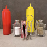 A condiment set on a gray surface: red ketchup and yellow mustard squeeze bottles, a chrome plated combination rack with glass salt and pepper shakers, a folded red-and-white striped napkin, and metal napkin tongs against a plain background.
