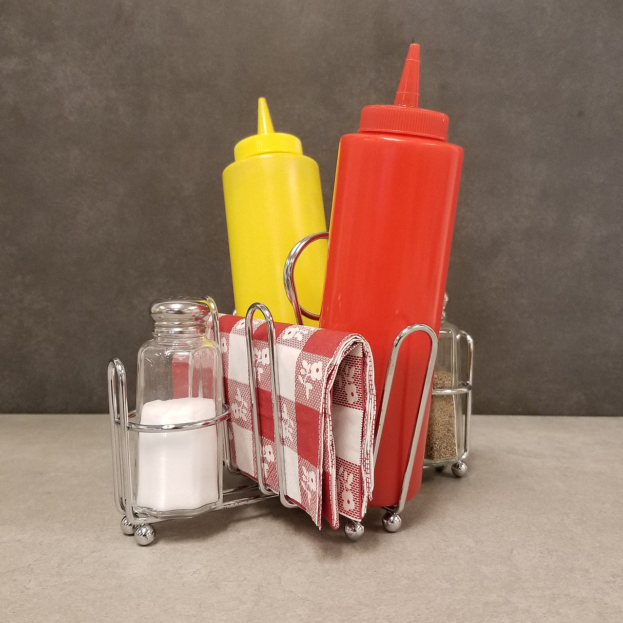 A chrome plated combination rack holds a yellow mustard bottle, a red ketchup bottle, salt and pepper shakers, and a stack of red and white checkered napkins against a plain gray background.