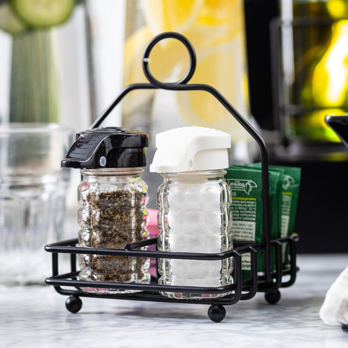 A black powder coated metal combination rack by TableCraft Products holds two glass shakers—one with black pepper, the other with salt—alongside green sugar packets. The caddy rests on a marble surface, with bottles and glassware blurred in the background.