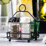A black powder coated metal combination rack by TableCraft Products holds two glass shakers—one with black pepper, the other with salt—alongside green sugar packets. The caddy rests on a marble surface, with bottles and glassware blurred in the background.