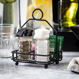 A black powder coated metal TableCraft Products combination rack holds a glass pepper shaker with a black lid, a glass salt shaker with a white lid, and two green tea packets on a marble countertop, glassware and lemon water pitcher in the blurred background.