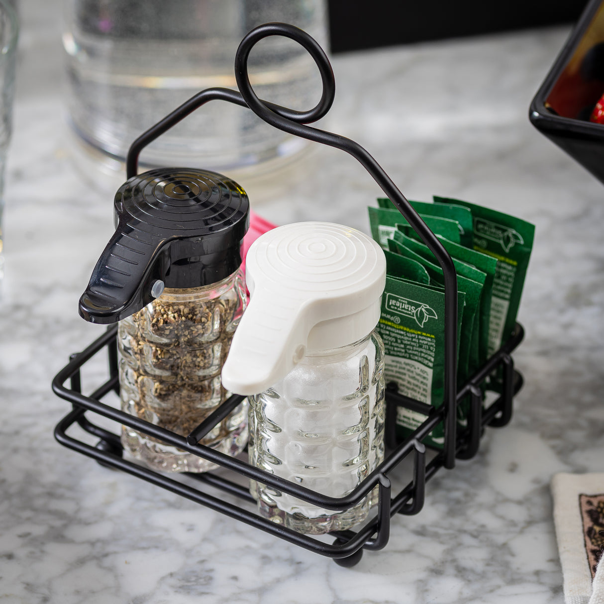 A TableCraft Products combination rack in black powder coated metal holds glass shakers—pepper with a black lid, salt with a white lid—and green sugar substitute packets on a marble tabletop, with a glass of water and part of a black bowl nearby.