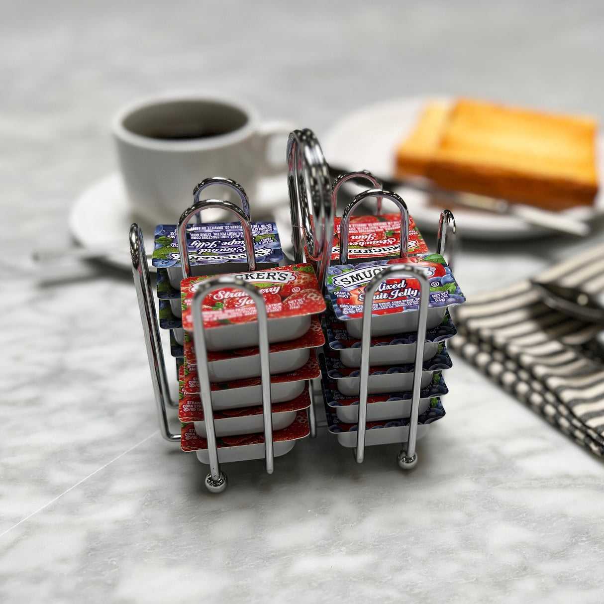 Two chrome plated metal racks hold individual jelly packet condiment packets of Smucker’s and similar jams on a marble table. In the blurred background are a cup of coffee, buttered toast, cutlery, and a folded striped napkin.