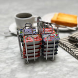 Two chrome plated metal racks hold individual jelly packet condiment packets of Smucker’s and similar jams on a marble table. In the blurred background are a cup of coffee, buttered toast, cutlery, and a folded striped napkin.