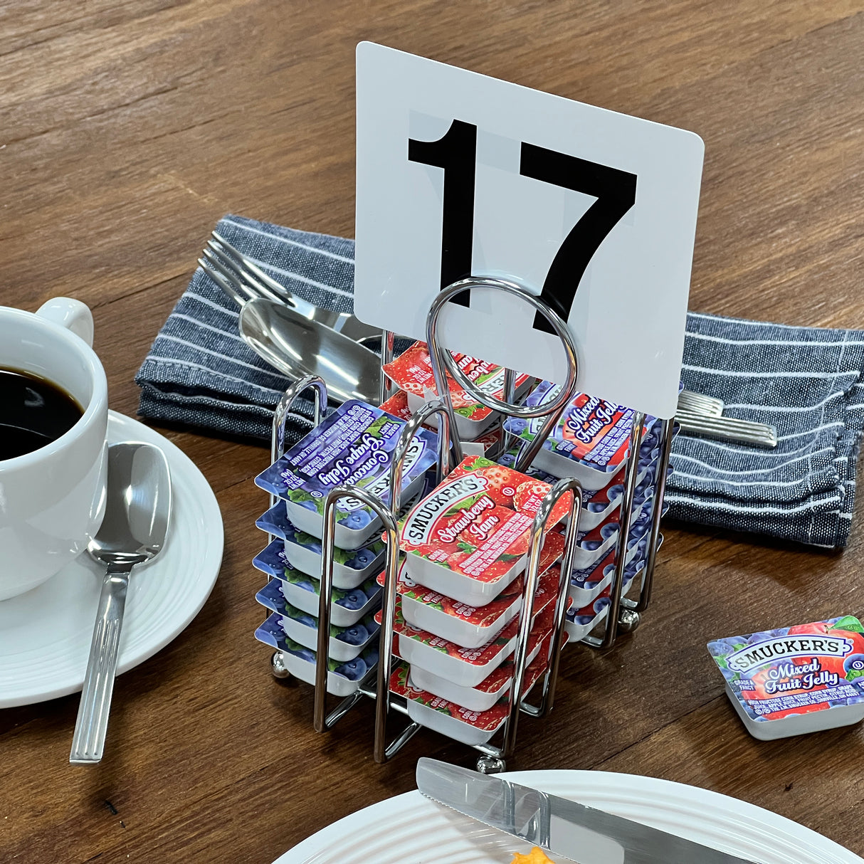 A wooden table displays a white cup of coffee, a plate, cutlery on a striped napkin, and a chrome plated jelly packet condiment rack with Smucker’s jelly packs and table number 17. A single jelly pack rests beside the caddy.