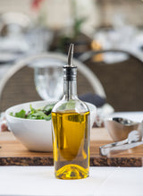 A clear glass bottle filled with yellow olive oil and topped with a stainless steel pourer sits on a white table. Behind it, there is a bowl of green salad, a metal garlic press, and blurred dining tableware in the background.