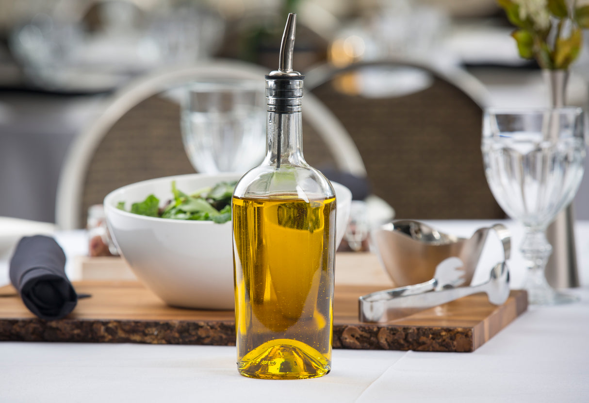 A glass bottle, fitted with a stainless steel pourer, sits on a table filled with golden olive oil. Behind it, a white salad bowl, metal tongs, black napkin, wooden board, and glassware await in an elegant dining setting.