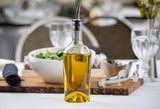 A glass bottle, fitted with a stainless steel pourer, sits on a table filled with golden olive oil. Behind it, a white salad bowl, metal tongs, black napkin, wooden board, and glassware await in an elegant dining setting.