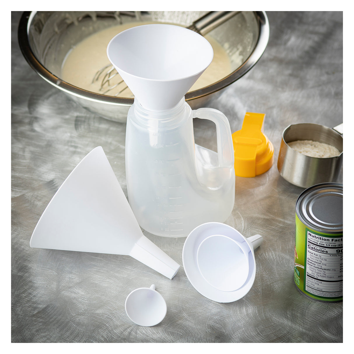 This plastic funnel set includes four white funnels of different sizes arranged on a grey countertop near a clear measuring jug. Dishwasher safe, they are perfect for kitchen tasks, with baking essentials and measuring cups in the background.
