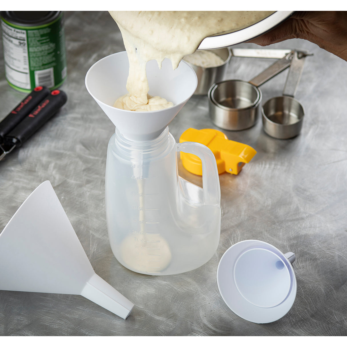 A hand pours thick batter from a bowl into a plastic container using a white funnel. Measuring cups, a yellow tool, a can, and part of a funnel set—dishwasher safe—are on the gray countertop, suggesting food preparation or baking.