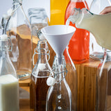 A hand pours a light-colored liquid from a jar into a glass bottle using a white plastic funnel. Surrounding the bottle are various dishwasher safe glass bottles filled with white, brown, and orange liquids, some with swing-top caps.