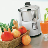 A shiny Waring 6001XK commercial juice extractor sits on a counter beside a basket of fresh produce, including carrots, apples, tomatoes, ginger, and leafy greens. A glass of bright orange juice is placed to the right of the juicer.