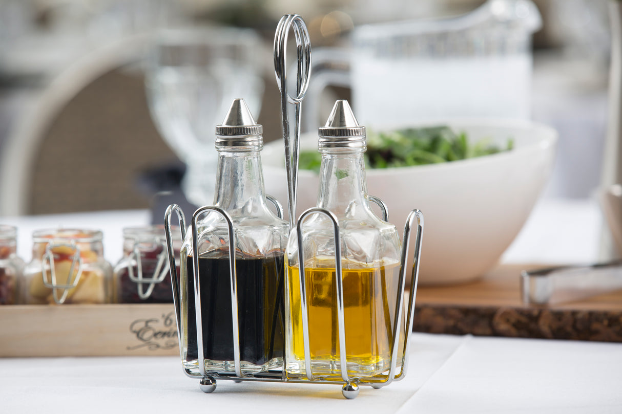 Two glass bottles with stainless steel tops rest in a metal Oil & Vinegar Dispenser on a white tablecloth. One holds dark vinegar, the other golden olive oil. A blurred bowl of greens and various small jars appear in the background.