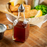 A glass oil & vinegar dispenser with a stainless steel top sits on a wooden table. In the blurred background, a basket lined with newspaper holds fried fish, lemon wedges, and green peas. A small sauce dish is partially visible to the left.