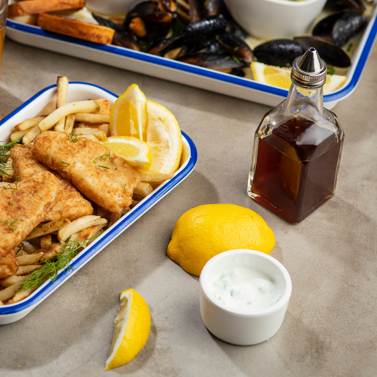 A tray of crispy fish and fries garnished with lemon wedges sits on the table. Beside it are a glass bottle oil & vinegar dispenser, a halved lemon, tartar sauce, and more lemon. A tray of mussels is partially visible in the background.