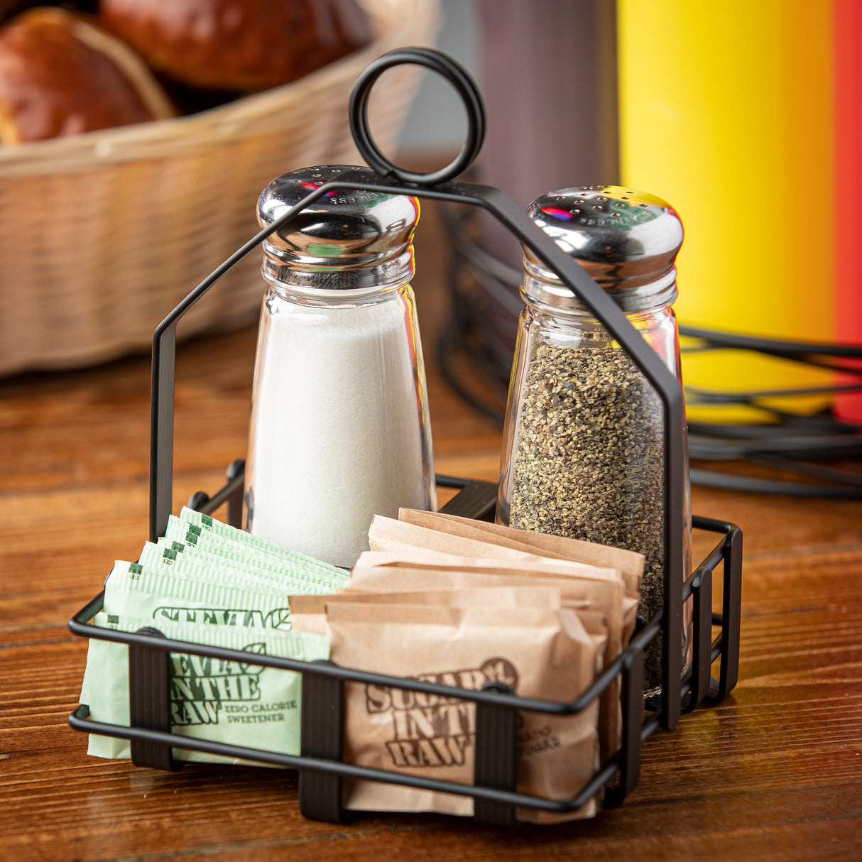 A black powder coated condiment rack holds glass salt and pepper shakers, brown “Sugar In The Raw” packets, and green zero-calorie sweetener on a wooden table. A basket of bread is blurred in the background.