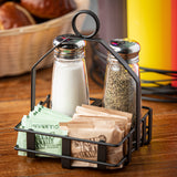 A black powder coated condiment rack holds glass salt and pepper shakers, brown “Sugar In The Raw” packets, and green zero-calorie sweetener on a wooden table. A basket of bread is blurred in the background.