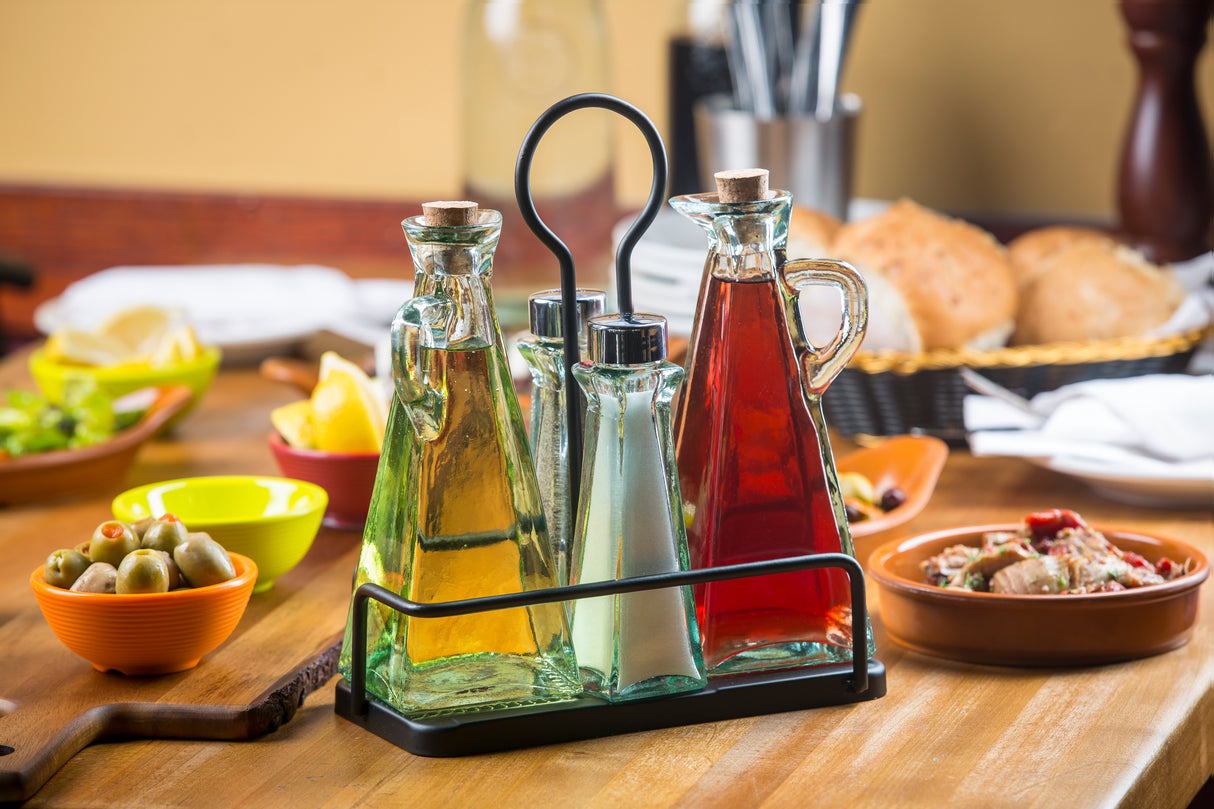 A TableCraft Marbella oil & vinegar set sits in a metal caddy on a wooden table, surrounded by bowls of olives, lemon wedges, salad, bread, and peppers—evoking a Mediterranean meal setting with cutlery in the background.